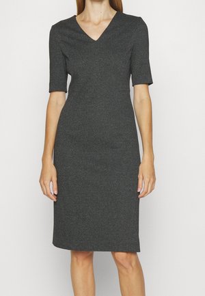 Femme portant une robe ajustée, longueur genou, gris foncé, à manches courtes et col en V, debout devant un fond clair uni.