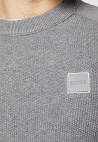 Sweat-shirt gris avec texture tricotée, encolure ronde et une étiquette rectangulaire gris clair avec "BOSS" embossé sur le côté supérieur gauche.