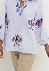 Světle modrá proužkovaná blůza s květinovou výšivkou v růžové a modré barvě, s výstřihem ve tvaru klíčové dírky a třásněmi, volné rukávy.