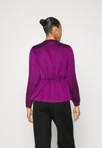 Blusa di satin viola con maniche a palloncino lunghe, vita stretta e texture liscia; indossata con pantaloni neri, vista da dietro.
