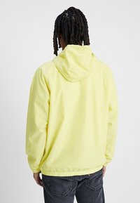 Gul huva jacka i lättviktsmaterial, med dragsko i huvan och elastiska ärmslut, i en avslappnad passform. Bakifrån visad.