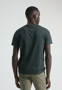 Camiseta de algodón verde oscuro con mangas cortas, cuello redondo, dobladillo recto y aberturas laterales, combinada con pantalones caqui claros. Textura lisa y sencilla.