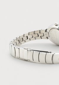 Ασημένιο ρολόι Calvin Klein από ανοξείδωτο ατσάλι με συνδεδεμένο βραχιόλι και μερικώς ανοιχτό κούμπωμα σε ουδέτερο φόντο.