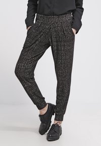 Czarne spodnie we wzory o luźnym kroju, z elastycznym paskiem w talii oraz ściągaczami u kostek, zestawione z czarnymi butami na sznurówki.