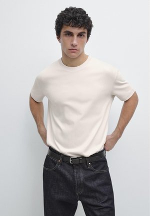 Junger Mann mit lockigem Haar, der ein schlichtes weißes Kurzarmhemd und dunkle Jeans trägt, steht mit den Händen in die Hüften gestützt vor einem schlichten Hintergrund.