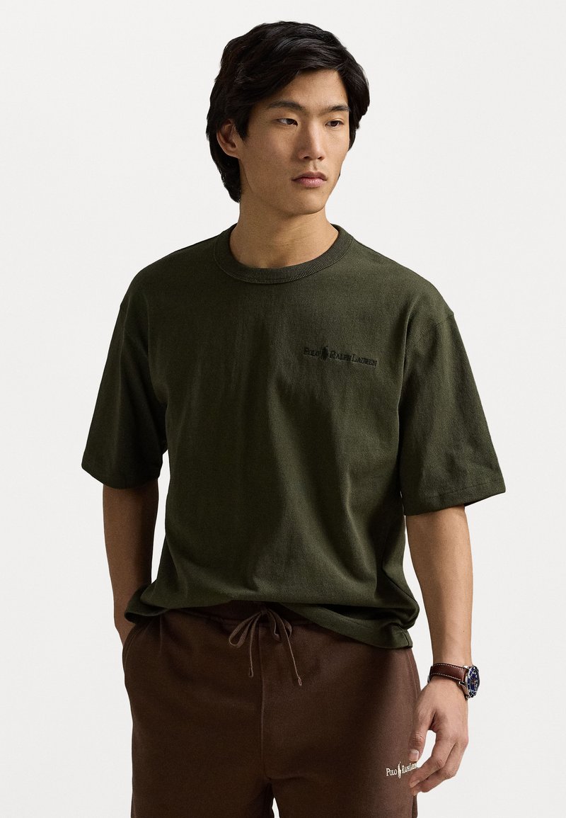 Tricou din bumbac de culoare verde oliv, cu gât rotund și mâneci scurte, având un mic logo negru în zona stângă a pieptului.