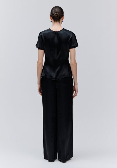 Frau mit zusammengebundenem Haar, trägt eine schwarze Satinbluse mit kurzen Ärmeln und weite Hosen, steht mit dem Rücken zur Kamera vor einem schlichten grauen Hintergrund.