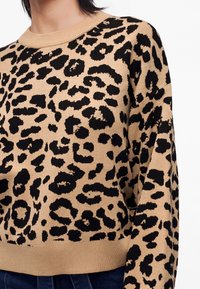 Žena má na sebe béžový sveter s čiernym leopardím vzorom a rebrovanými manžetami a spodným lemom, kombinovaný s tmavomodrými nohavicami.