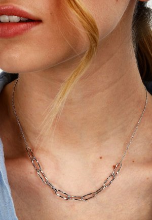 Nahaufnahme des Nackens einer Person, die eine silberne Kettenhalskette mit großen rechteckigen Gliedern trägt, während eine Strähne helles Haar über den Nacken fällt.