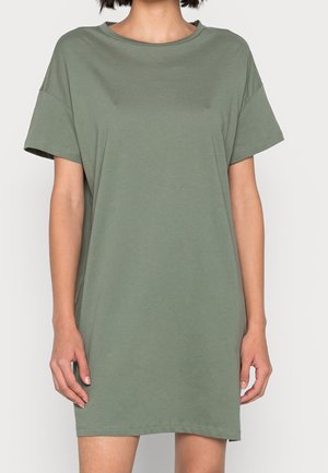 Vrouw draagt een olijfgroene T-shirtjurk met korte mouwen en losse pasvorm, staande tegen een effen lichte achtergrond.
