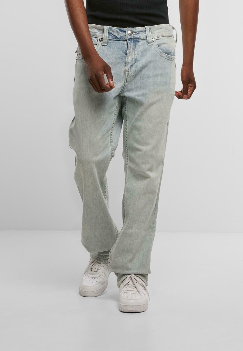 Heledad sinised denim teksad, vabalt istuvad, koos nööbiga sulgemise, viie tasku ja kergelt pleekinud välimusega, kantud koos valgete tennisereitega.