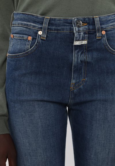 Jeans de mezclilla azul oscuro con cintura alta, herrajes de cobre, cinco bolsillos y detalles de costura en contraste. Textura suave en todo.