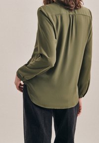 Blusa verde oliva con maniche lunghe, tessuto morbido, leggero sheen e orlo arrotondato, con schiena plissettata e polsini con bottoni.