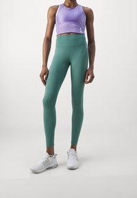 Ljuslila crop top med smala axelband, kombinerad med högmidjade, åtsittande leggings i grönt. Bärs med vita sportskor.