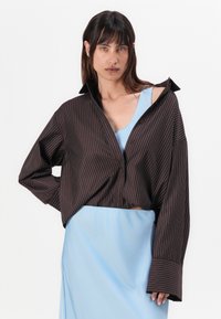 Chemise oversize à rayures brunes avec un col, associée à une robe de nuit en satin bleu clair. Le tissu semble lisse ; la chemise a des manches longues amples.