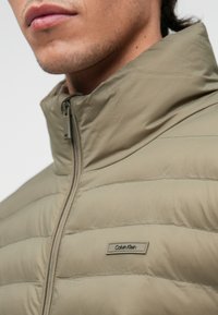 Šviesiai ruda pūkinė striukė su aukštu apykakle, vertikaliomis siūlėmis ir šoniniu užtrauktuku. Ant krūtinės yra prekės ženklo etiketė, skirta identifikacijai.