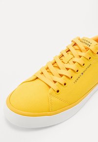 Pantofi sport din pânză galbenă cu o talpă din cauciuc albă, având șireturi plate galbene, un design cusut și un logo "Tommy Hilfiger" pe partea laterală.