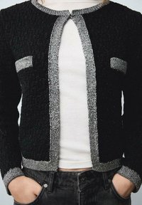 Cardigan noir texturé avec bordure argentée, encolure ronde et poches avant. Présente un design légèrement structuré et des manches ajustées.