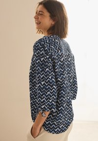 Blusa blu navy con motivo a zigzag in tonalità più chiare. Maniche a tre quarti con lacci ai polsini. Tessuto leggero e morbido.