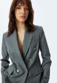 Grijze dubbelzijdige blazer met metalen knoopaccenten, notch-lapels en voorzakken. Textuurstof met een getailleerde pasvorm.