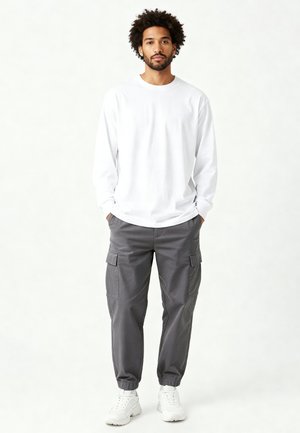 Giovane uomo con capelli ricci che indossa una maglia bianca a maniche lunghe, pantaloni cargo grigi e sneakers bianche, in piedi con le mani nelle tasche.