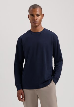 Navyblaues Langarmshirt aus glattem Baumwollmaterial, mit Rundhalsausschnitt und dezentem Logo auf der Brust. Stilisiert mit beigen Hosen.