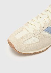 Light beige sneaker with a textured suede upper, mesh panels, and a light blue accent. Features soft laces and a rubber sole.