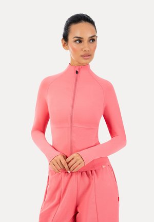 Frau trägt eine eng anliegende, langärmelige pinke Reißverschlussjacke und passende pinke Hose, schaut leicht zur Seite vor einfachem Hintergrund.