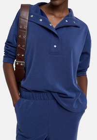 Granatowy sweter z klapkowym kołnierzem, zatrzaskami oraz luźnym krojem. Posiada miękką teksturę i elastyczny pas. Brązowy pasek na ramieniu.