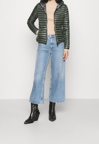 Zelená péřová bunda se zipem vpředu, pruhovaná textura, kombinovaná se světle modrými širokými džínami a černými špičatými kotníkovými botami.