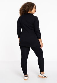 Zwarte tuniek met lange mouwen, gecombineerd met zwarte leggings en witte sneakers met zwarte strepen. Glad materiaal, relaxte pasvorm.