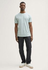 Camiseta de manga corta verde claro con cuello redondo, combinada con jeans azul oscuro y zapatillas blancas con detalles en beige y suelas texturizadas.