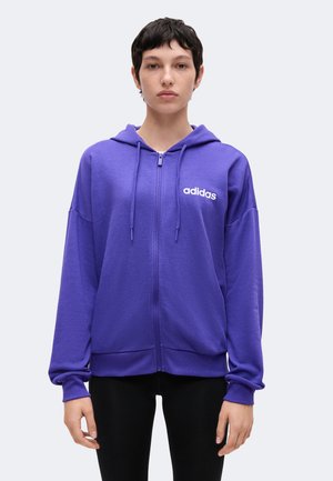 Osoba nosząca fioletową bluzę Adidas na zamek z kapturem i sznurkami, stojąca prosto na jednolitym jasnym tle.