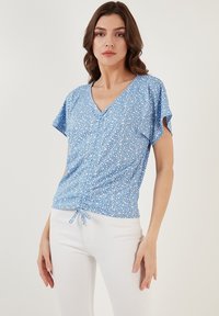 Blusa a fiori blu con scollatura a V, maniche corte e dettaglio arricciato con legatura in vita. Tessuto leggero e texture.