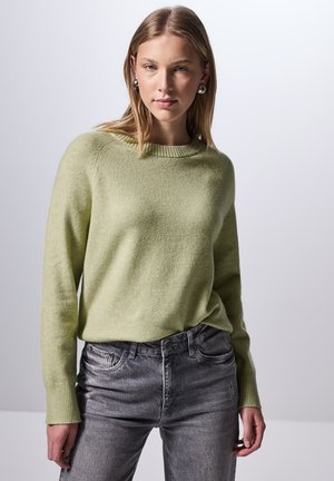 Pull en maille vert clair avec un col rond et des poignets côtelés, associé à un jean gris délavé. Texture douce avec une coupe décontractée.