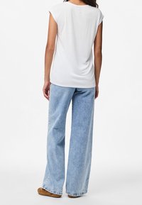 Person klädd i en vit ärmlös topp, ljusblå vida jeans och bruna skor, stående med ryggen mot betraktaren på vit bakgrund.