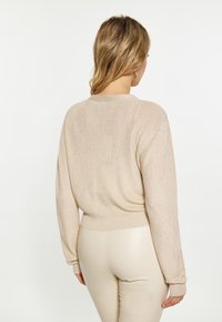 Maglione lavorato a maglia beige con maniche lunghe, polsini a costine e dettaglio sul retro. Abbinato a pantaloni beige aderenti, che mostrano una morbida texture e lucentezza.