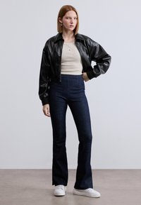 Μαύρο μπουφάν από συνθετικό δέρμα με cropped σχεδιασμό, συνδυασμένο με ψηλόμεσα σκούρα τζιν παντελόνια με καμπάνα και λευκά αθλητικά παπούτσια.