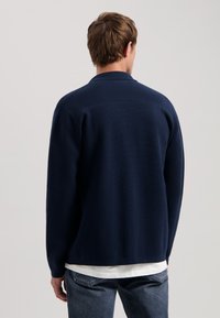 Námornícky modrý pletený cardigan s golierom a dlhými rukávmi, s textúrovaným povrchom a rovnými línie, nosený cez bielu košeľu.