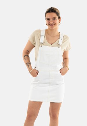 Vestido de tirantes en denim blanco con correas ajustables, bolsillos frontales y una silueta ajustada, combinado con una camiseta de color beige claro.