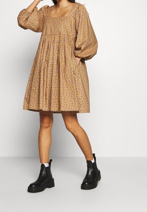 Frau trägt ein locker sitzendes, knielanges, senffarbenes Kleid mit blauen Blumenmustern und Puffärmeln, kombiniert mit schwarzen Stiefeletten und weißen Socken.