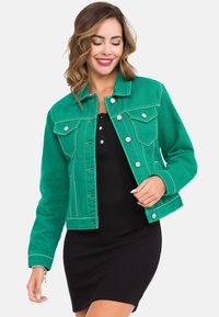 Grüne Denimjacke mit einem Kragen, vorderer Knopfverschluss, zwei Brusttaschen und sichtbaren Steppnähten. Über einem schwarzen Kleid getragen.