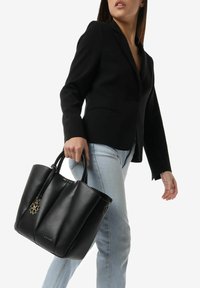 Zwarte leren tote bag met zijsplitsingen, platte handvatten en een decoratieve charm. De tas heeft een gestructureerde vorm en een minimalistisch ontwerp.