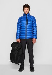 Doudoune bleue avec texture matelassée et fermeture éclair à l'avant ; sac à dos noir tenu dans la main gauche ; pantalon et chaussures noirs ; arrière-plan minimal.