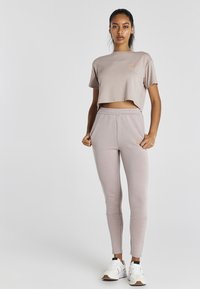 Vaalean taupe-väriinen lyhythihainen t-paita ja siihen sopivat kapenevat housut, jotka on valmistettu pehmeästä kankaasta. Vaatekaappia koristaa brodeerattu logo ja sivutaskut, joita on yhdistetty valkoisiin lenkkareihin.