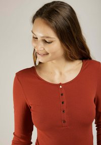 Haut à manches longues couleur rouille avec un décolleté en scoop et patte de boutonnage à cinq boutons. Présente une texture côtelée et une coupe ajustée.