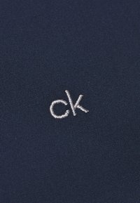Tejido marino con una textura fina que presenta un logo "ck" bordado en plata, situado de manera destacada en la superficie.