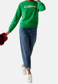 Felpa verde con testo "California", abbinata a jeans denim blu, leggermente morbidi, e sneakers bianche; berretto rosso tenuto in mano.