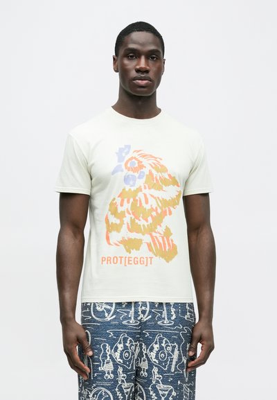 Ανοιχτό πράσινο T-shirt με κοντά μανίκια, που απεικονίζει ένα πολύχρωμο πουλί και το κείμενο "PROT[egg]T" σε πορτοκαλί στο κάτω μέρος. Μαλακό βαμβακερό υλικό.