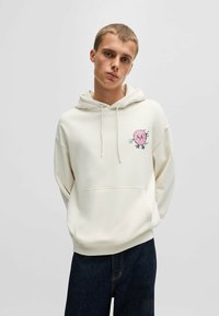 Hoodie beige clair en tissu doux avec une poche avant, arborant un graphique de donut cartoon rose sur la poitrine gauche. Capuche ajustable avec cordon.